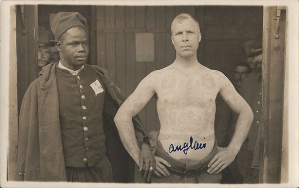 Afrikanischer und englischer Kriegsgefangener. Rückseite: „Emil Meiner, Foto Atelier, Ohrdruf i./Thür“. Ansichtskarte, ungelaufen, ohne Datum. Sammlung Detlev Brum. Die Zuordnung zu Ohrdruf ergibt sich allein aus der Verlagsangabe. Möglich ist aber auch, dass das Bild in einem Kriegsgefangenenlager in Münster entstanden ist und (rechts) einen englischer "Boxer" ablichtet. Die Hand- bzw. Armreichungsszene dürfte eine vom Fotograf in Szene gesetzte homoerotische Komponente signalisieren, die die englisch-afrikanische "Verbrüderung" karikieren soll.