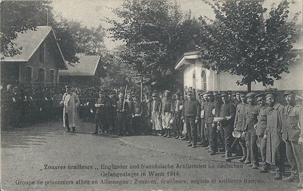 "Zouaves trialleurs, Engländer und französische Artilleristen im deutschen Gefangenenlager in Wahn 1914". Carte Postale, ungelaufen. "N. Sch. Brux. – Reprod. Interd. – Autor. Par le gouv. Allemand, Brux." Sammlung Detlev Brum. Die zweisprachige Version ist vermutlich eine Ansichtskarte, die speziell für Kriegsgefangene angefertigt wurde, die solche Ansichtskarten in die Heimat verschickten. Eine bildidentische, jedoch s/w-Ansichtskarte (Verlag Carl Rud. Bremer & Co, Köln a. Rhein) mit dem Titel "Im Gefangenenlager. Antreten zum Essenholen" dürfte für den Versand in Deutschland genutzt worden sein.